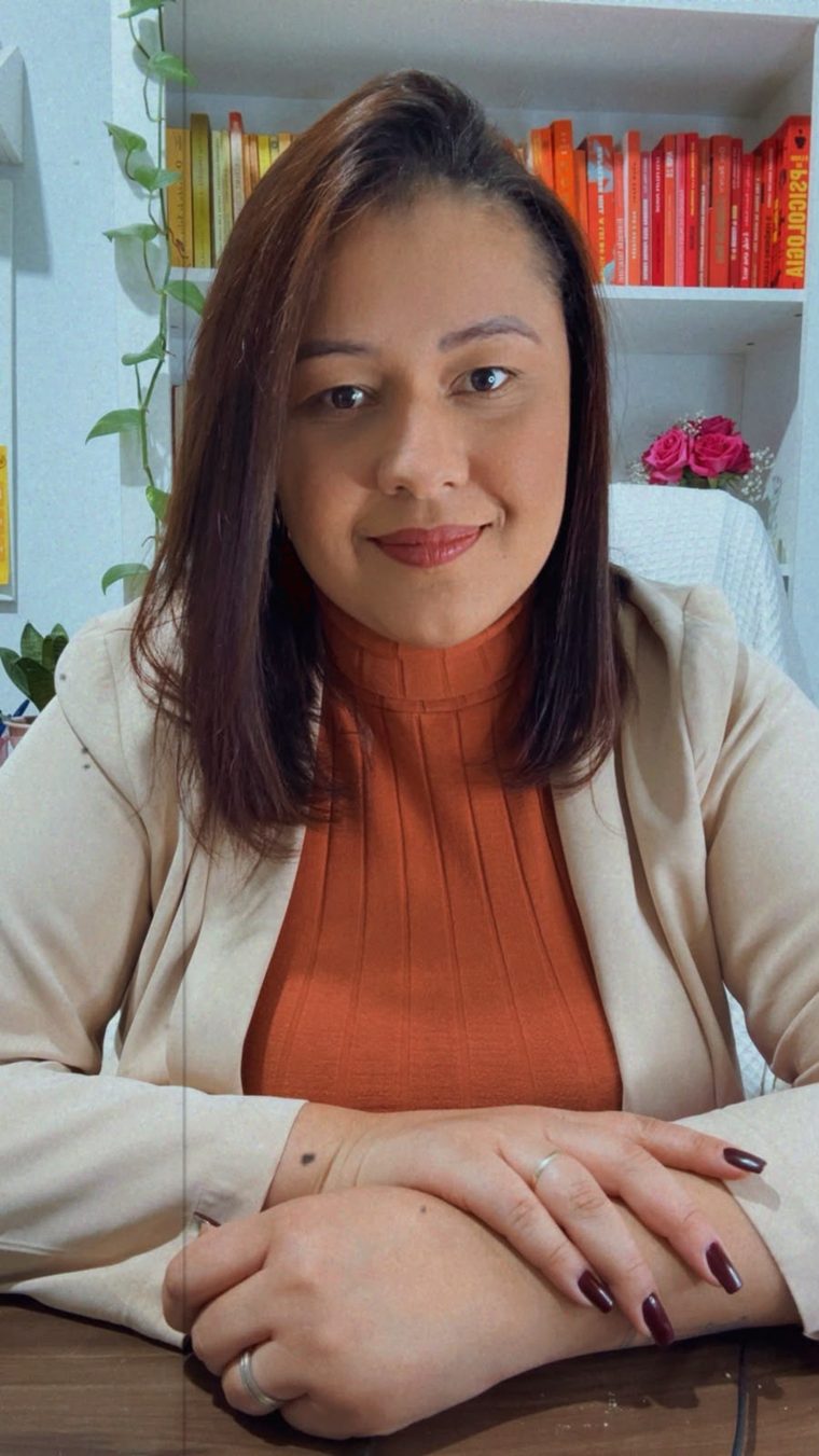 Karla Karolina, psicóloga, de blazer bege, sorrindo, em frente a uma estante de livros